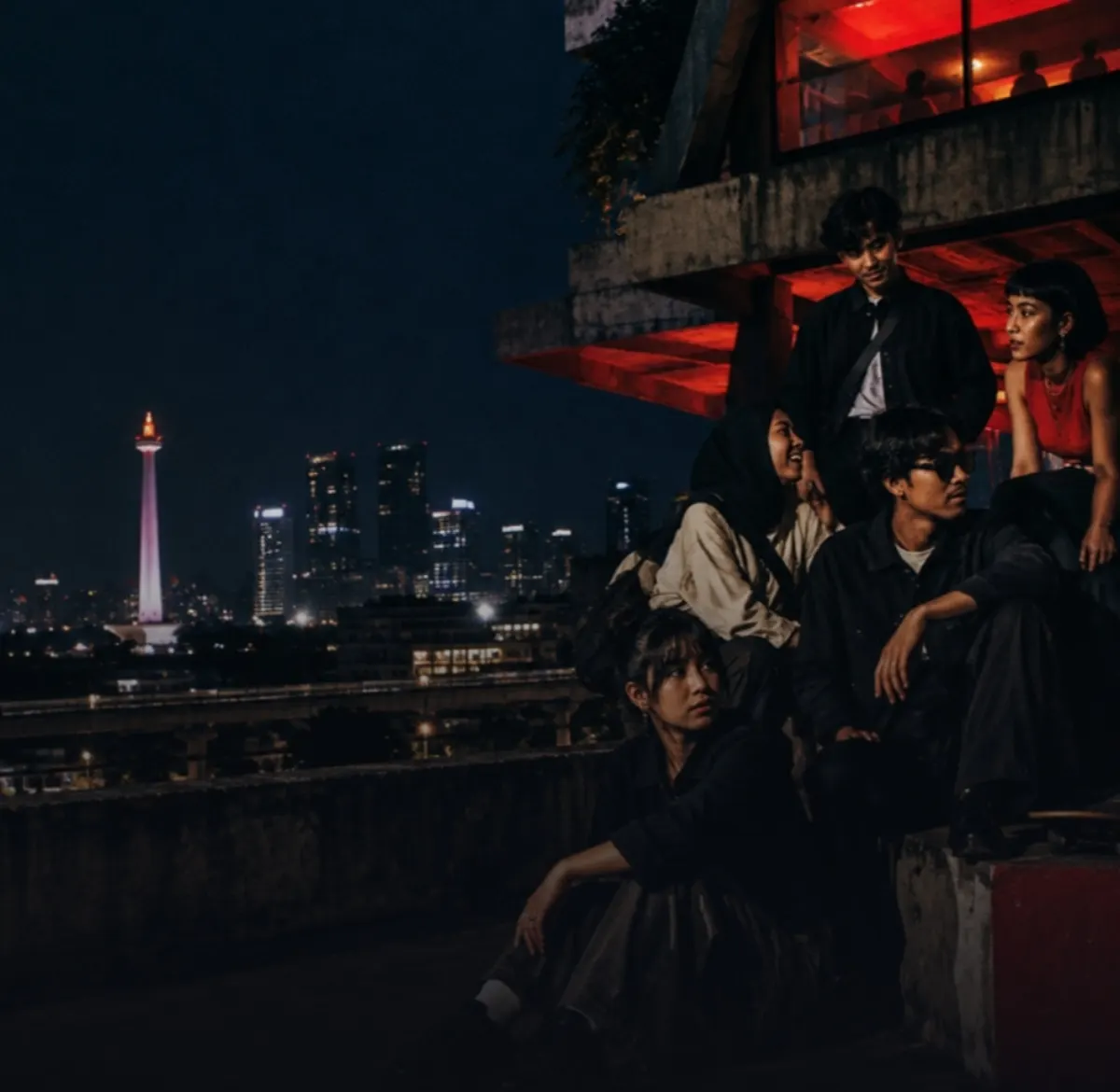 Young people gathered on a Jakarta rooftop at night, with the Monas monument lit up behind them
